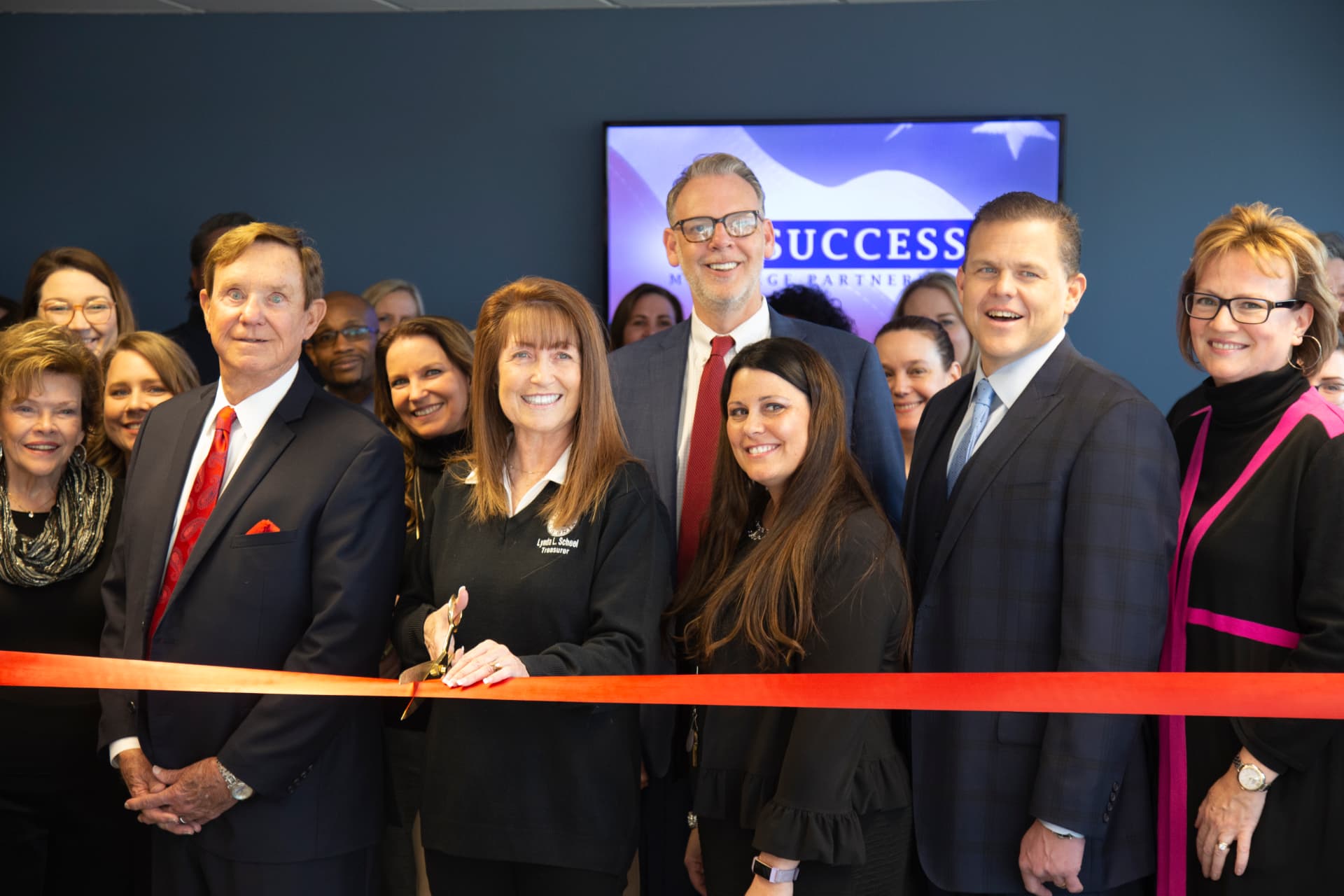 Success Mortgage Partners team members at a ribbon-cutting ceremony, a woman at the center cutting a red ribbon with gold scissors