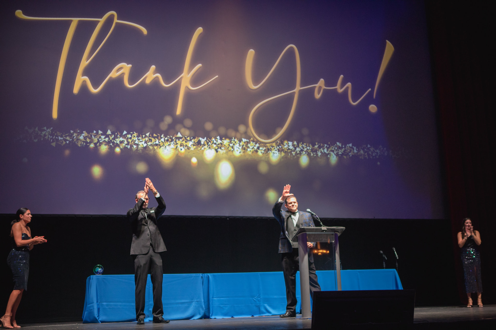 Owen Lee at a lectern on a gala stage, a large screen behind him reading Thank You! in gold script, colleagues applauding on either side